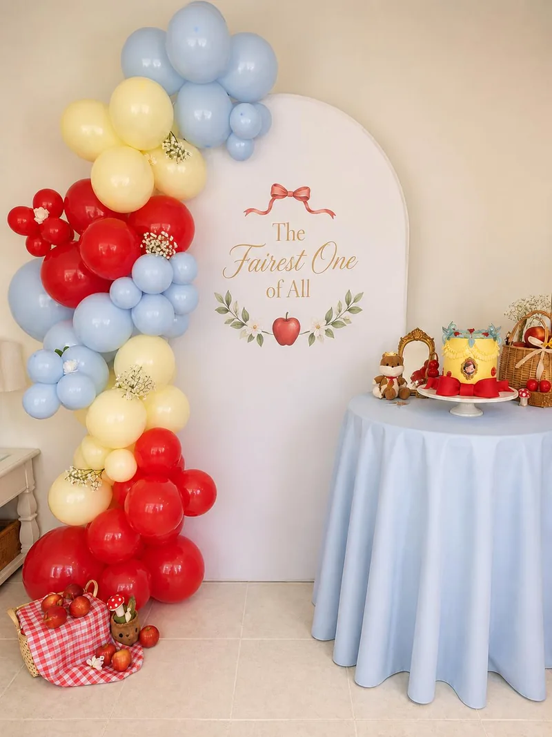 Red, pale yellow and blue balloons around a white fairytale backdrop with apple props and a cake table.