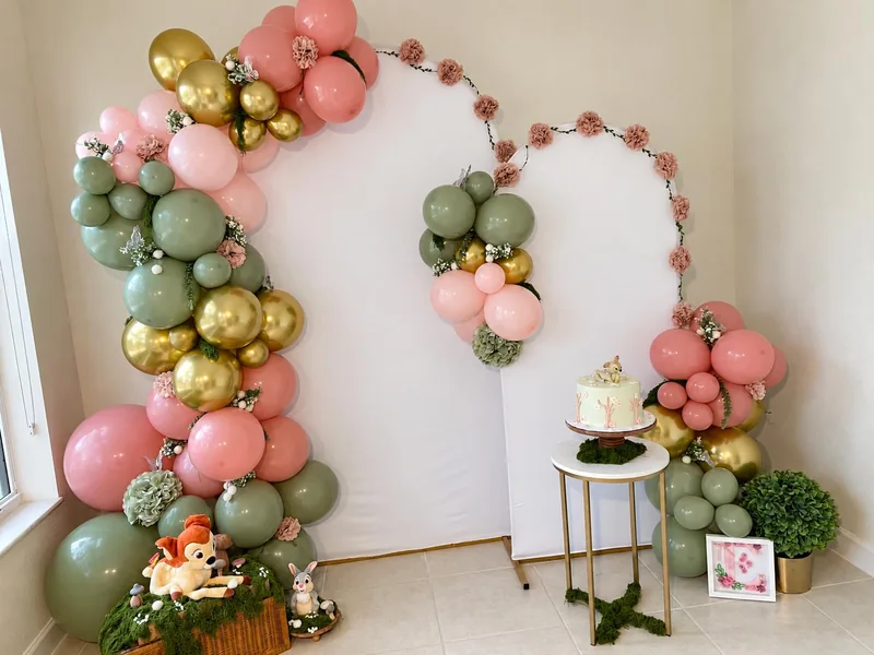 Pink, sage and gold balloon garlands around white arch backdrops with woodland props and a cake pedestal.